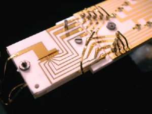 NIST physicists demonstrated sustained, reliable quantum information processing in the ion trap at the left center of this photograph, improving prospects for building a practical quantum computer. The ions are trapped inside the dark slit (3.5mm long and 200 micrometers wide) between the gold-covered alumina wafers. By changing the voltages applied to each of the gold electrodes, scientists can move the ions between the six zones of the trap.
Credit: J. Jost/NIST