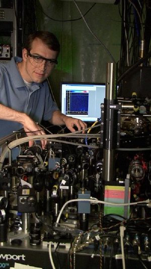 NIST postdoctoral researcher David Hanneke at the laser table used to demonstrate the first universal programmable processor for a potential quantum computer. A pair of beryllium ions (charged atoms) that hold information in the processor are trapped inside the cylinder at the lower right. A colorized image of the two ions is displayed on the monitor in the background. Credit: J. Burrus/NIST
