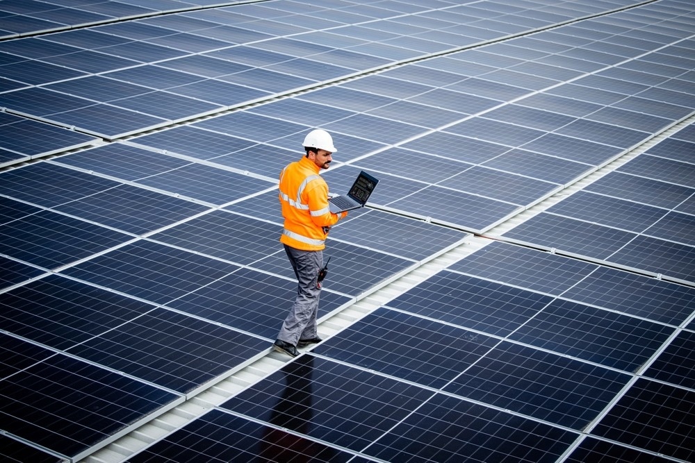 Renewable energy engineer walking through large solar power plant and checking electricity production on his laptop computer.