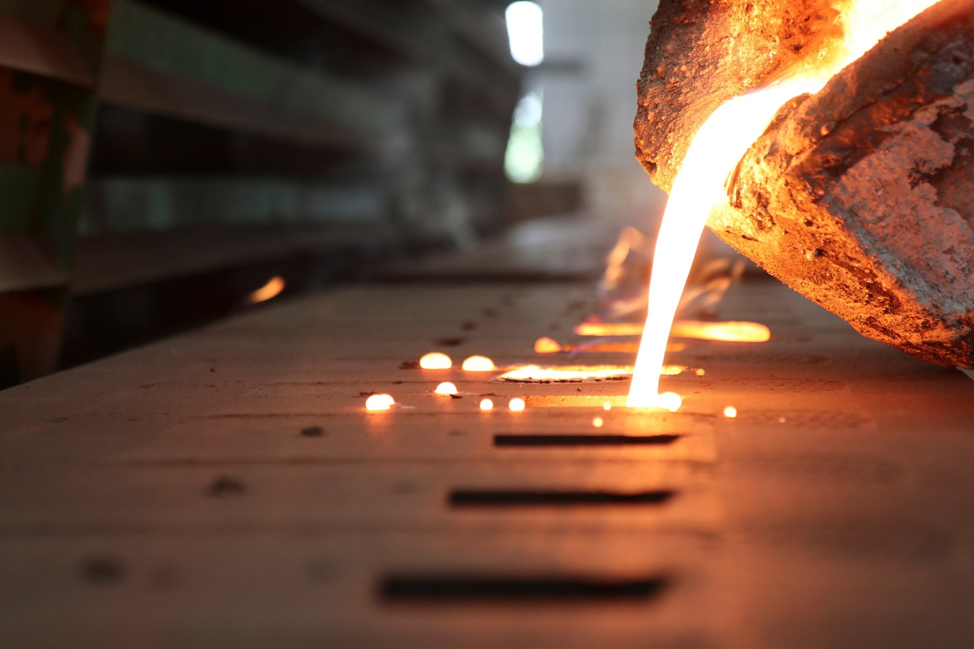 Molten metal being poured into molds on a flat surface in a casting facility. Bright orange molten metal streams from a ladle into rectangular molds, with sparks and