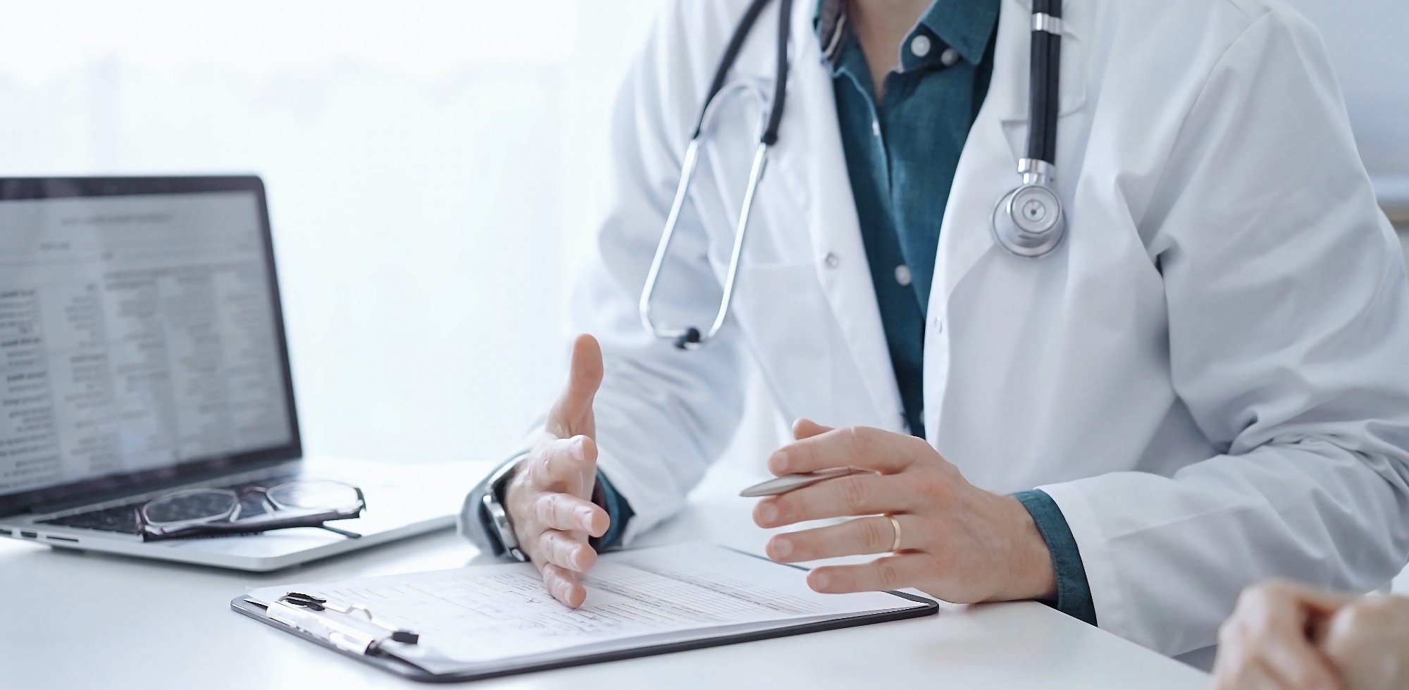 Doctor and a patient. The physician, wearing a white medical coat over a green shirt, is gesturing with his hands during a consultation in the clinic.