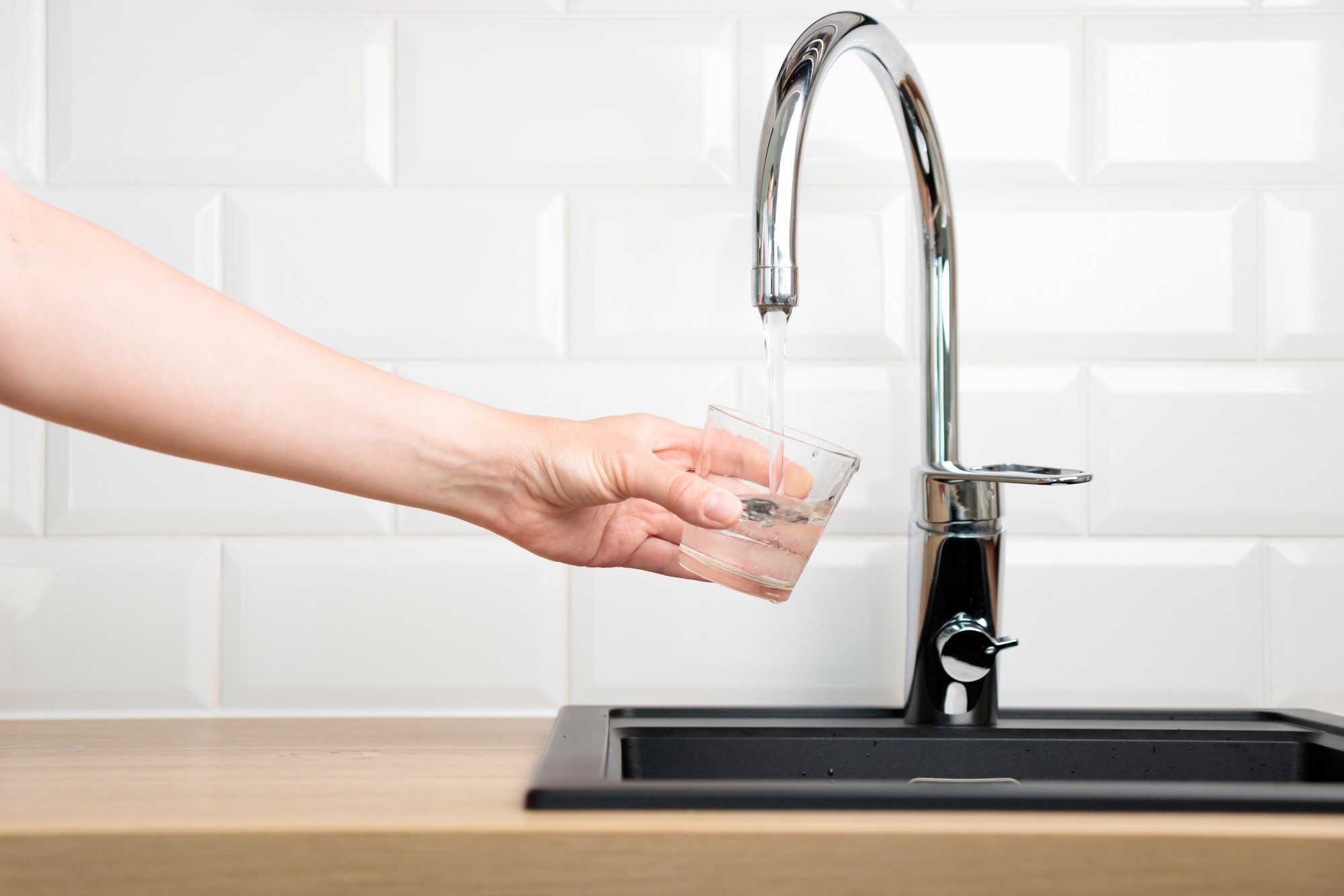 A hand holding a glass under a modern kitchen faucet, with water being poured into the glass from the faucet. The background shows a white tiled wall and a dark kitchen sink.