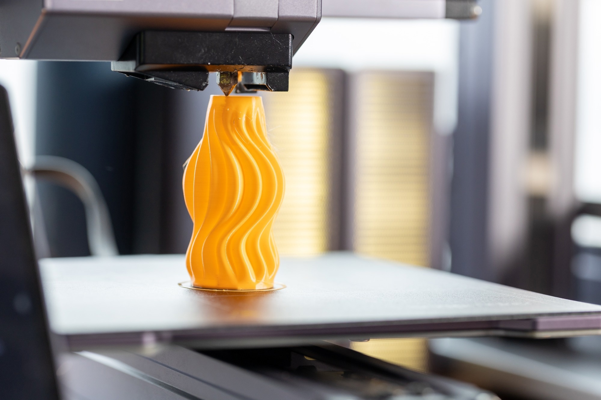 A 3D printer creating a yellow, swirling shape with a smooth surface. The printer nozzle is extruding the material as it builds the object layer by layer.