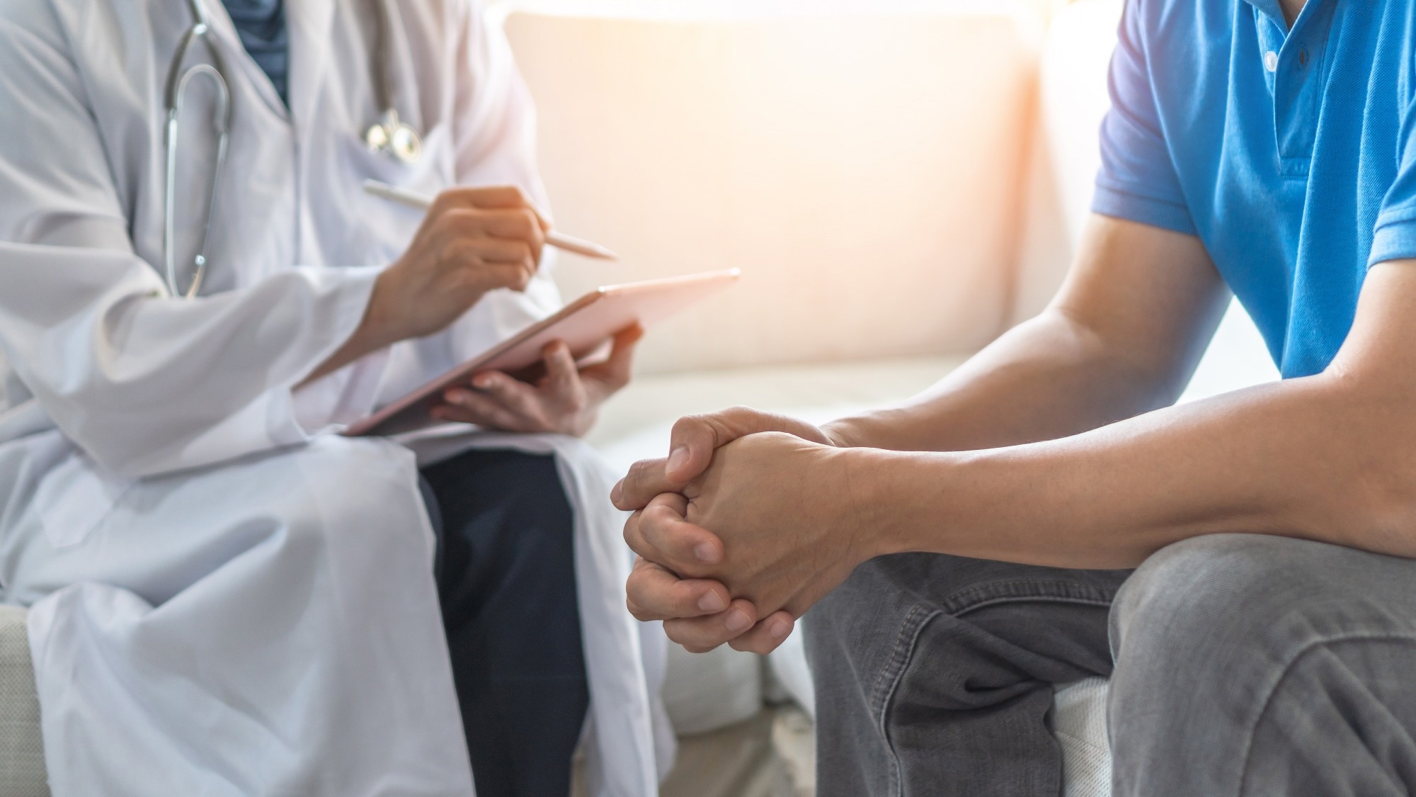 Close-up of a doctor in a white coat holding a digital tablet while consulting with a patient. The patient, seated and wearing a blue shirt, clasps their hands together. They are having a serious or attentive conversation in a clinical setting.