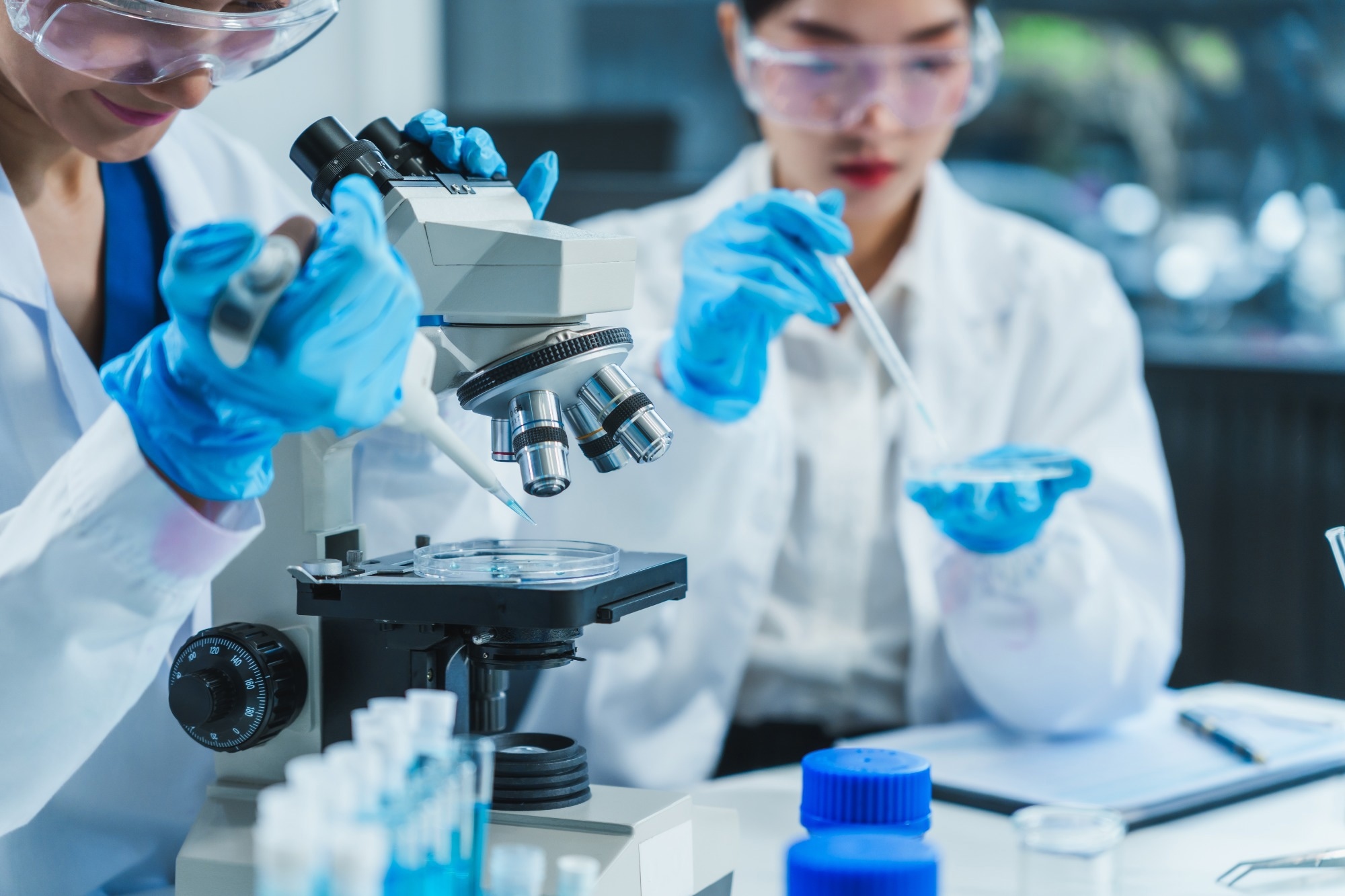 Two women in a lab work on food research, using a microscope, petri dish, and test tubes filled with chemical solutions.