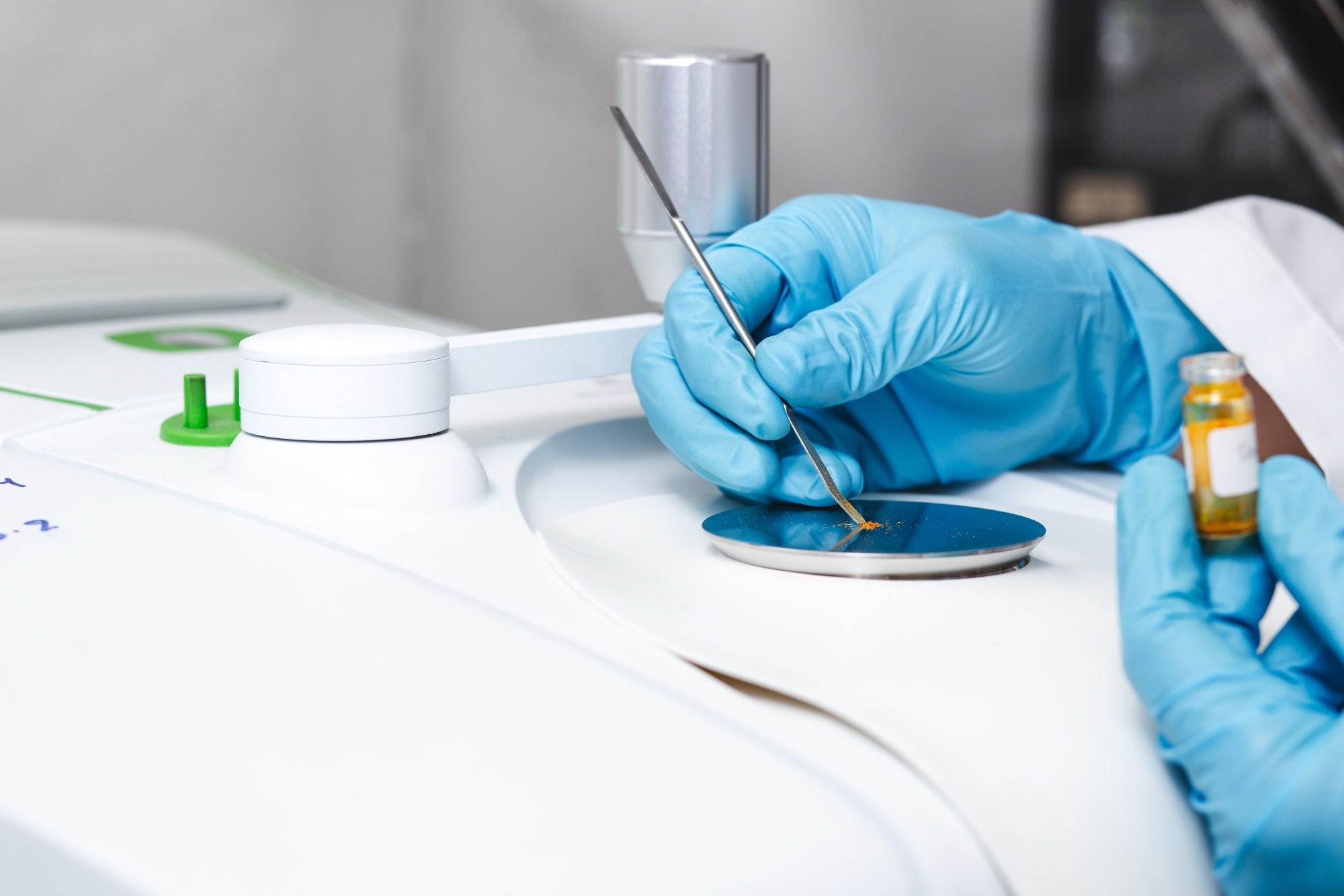 A scientist wearing blue gloves placing a yellow powder onto the sample plate of an FTIR spectrometer.