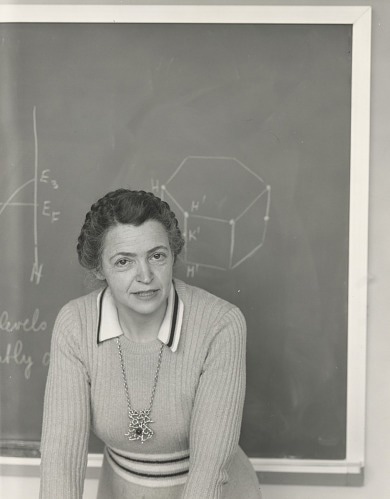 Millie stands in front of a black chalkboard with scientific equations on it. She is leaning forward and looking into the camera. (Image usage rights granted by Schlesinger Library)