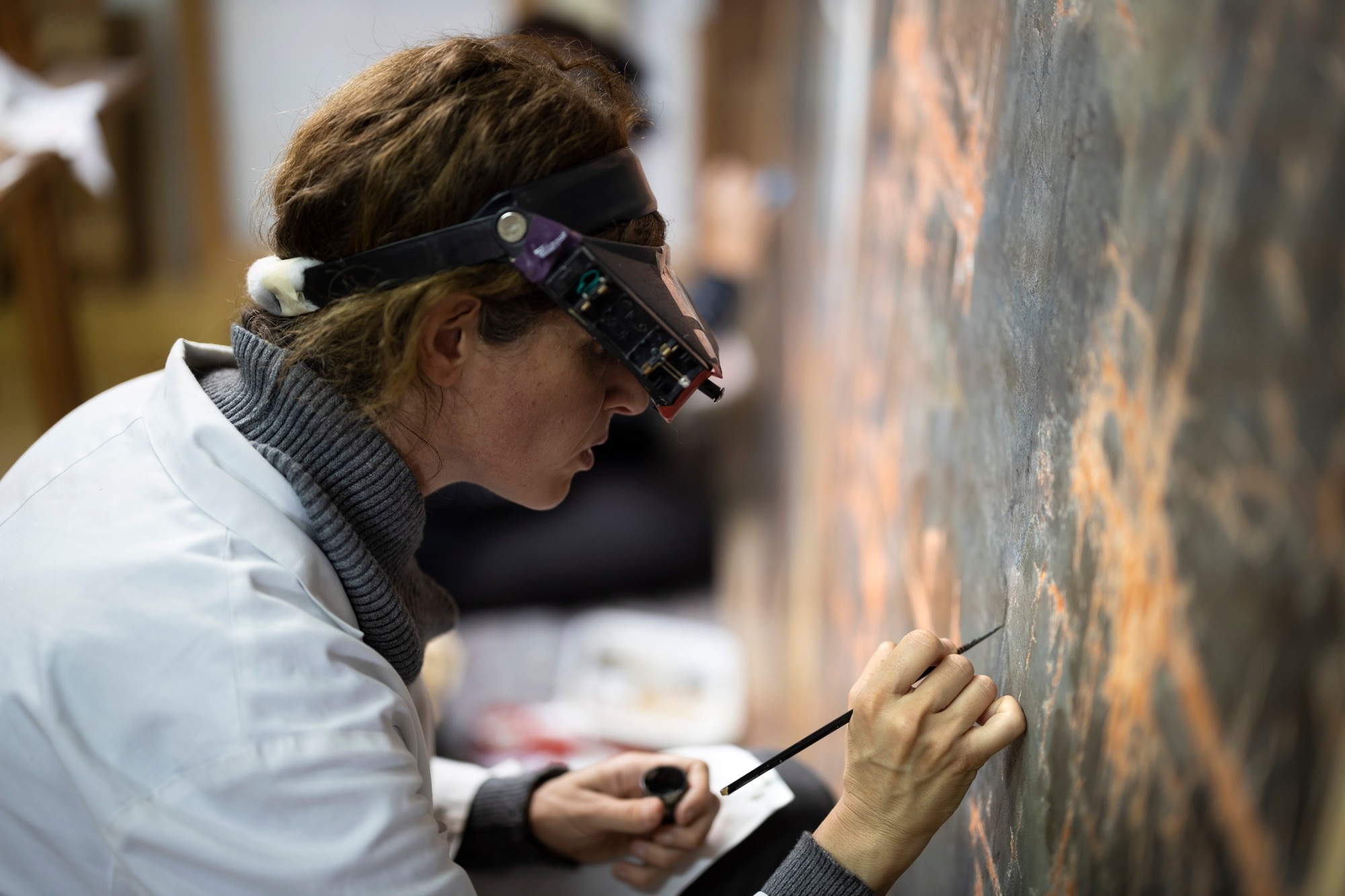 An art restorer gently applies paint to a painting she is restoring. She wears a lab coat and googles.