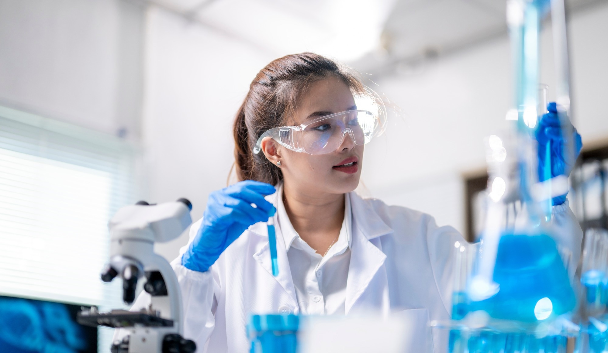 A woman in a lab coat is holding a test tube and wearing safety goggles. She is in a lab setting, surrounded by various scientific equipment and glassware.
