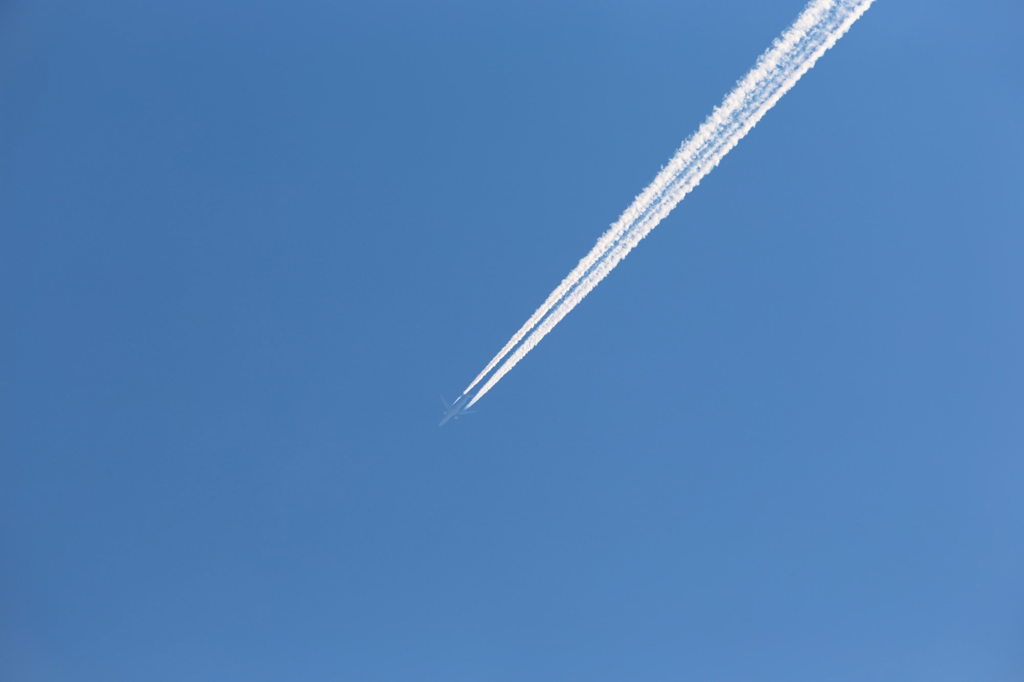 An almost invisible plane flies across a blue sky, leaving bright white contrails in its path.