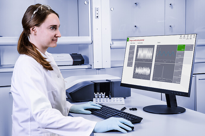 A scientist (woman wearing lab coat, nitrile gloves and lab goggles) sits at a desk and reviews spectra from the Litesizer on a desktop.