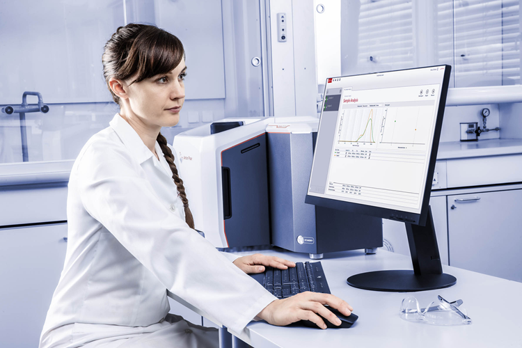 A scientist reviews spectra from the analyzer on a computer screen. She wears a labcoat and has dark hair and brown eyes.