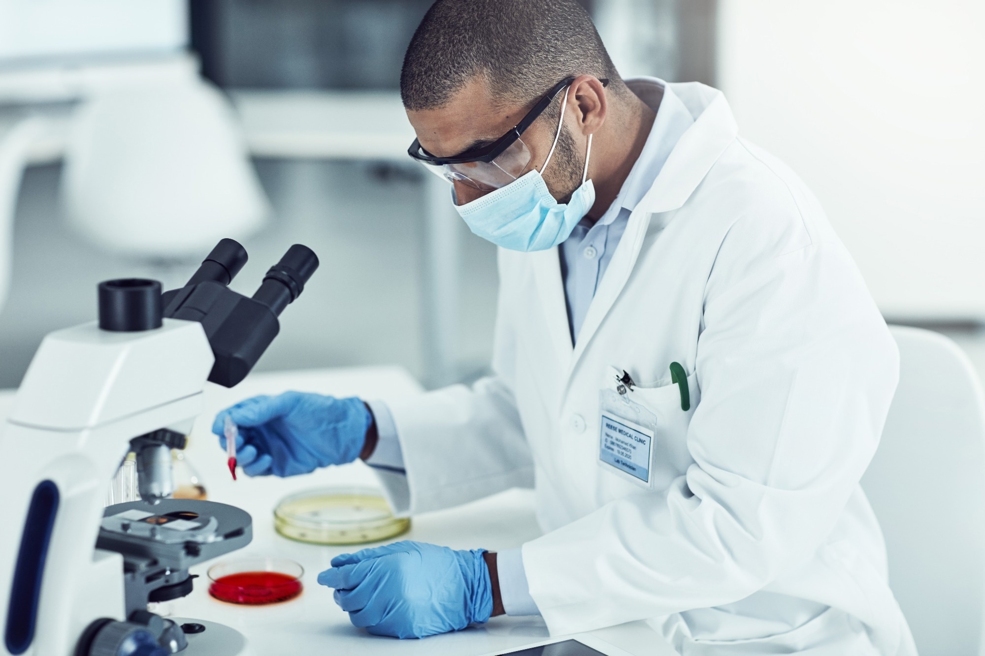 A scientist wearing protective glasses, a face mask, and gloves, working in a laboratory with a microscope and handling a sample in a small vial.
