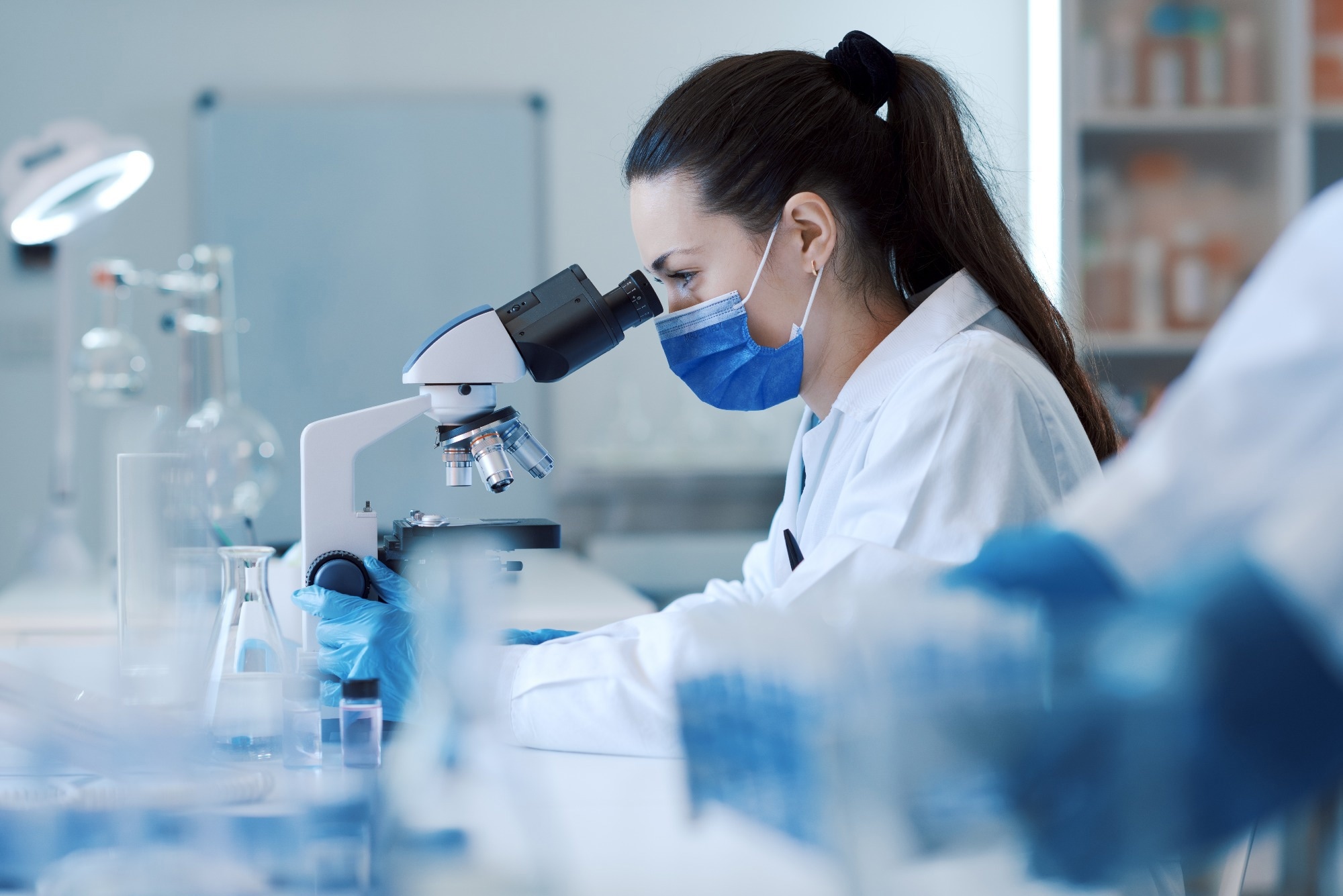 Female scientist wearing a protective mask and gloves, examining samples under a microscope in a laboratory