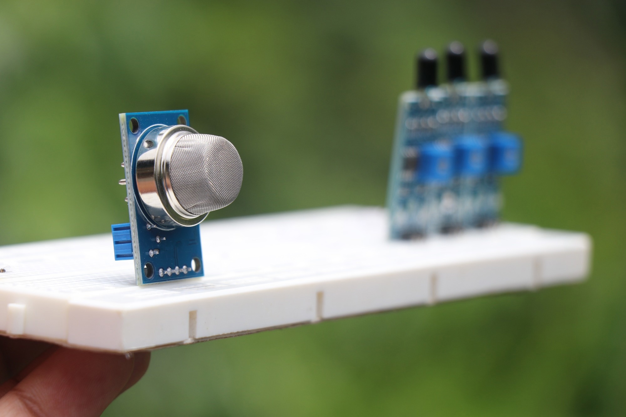 Close-up of an electronic gas sensor module mounted on a prototype board, featuring a cylindrical metal mesh component for gas detection, set against a blurred green background.
