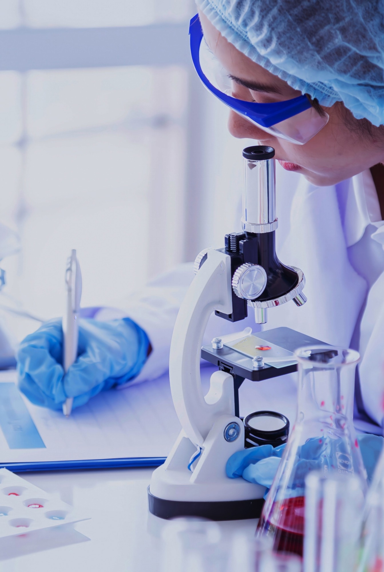 Scientist in protective gear using a compound microscope and taking notes in a laboratory, surrounded by glassware and lab tools