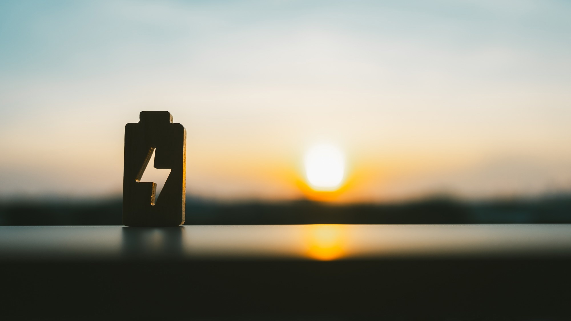 Silhouette of a battery-shaped icon with a lightning bolt cutout placed on a ledge, with a bright sunset in the background