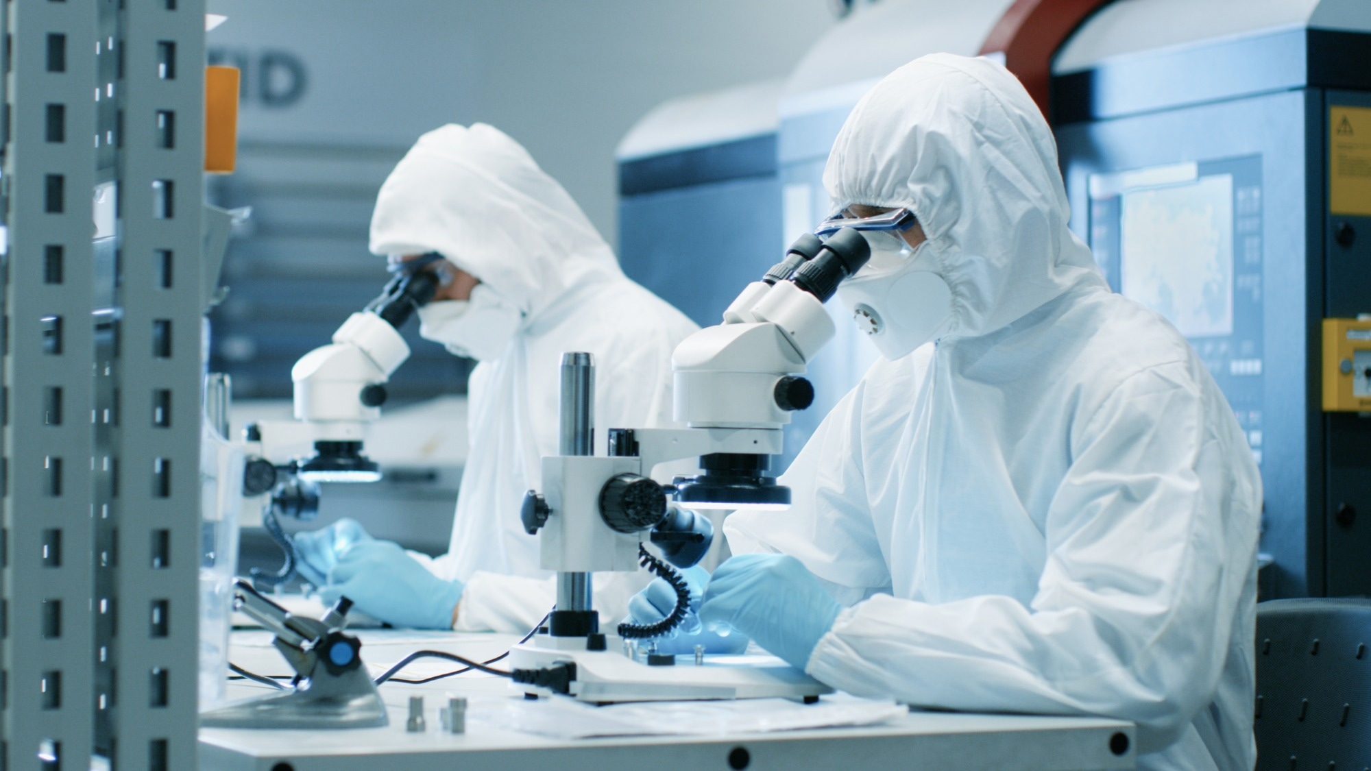 Two scientists in an ultra clean lab examining a material under a microscope.